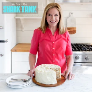 Woman in a pink shirt presenting a cake in a modern kitchen.